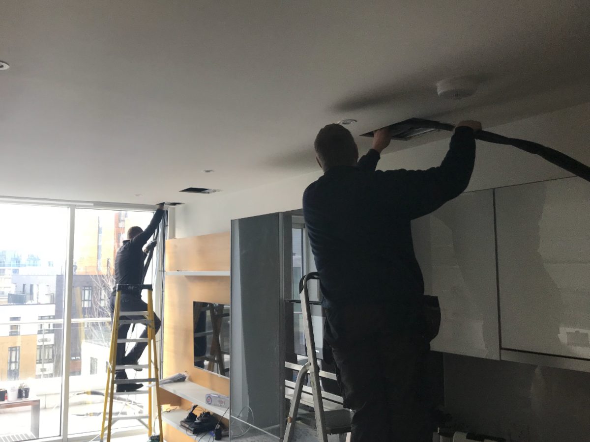 Two fitters on ladders feeding pre-insulated pipe into cut ceiling slots in a finished apartment