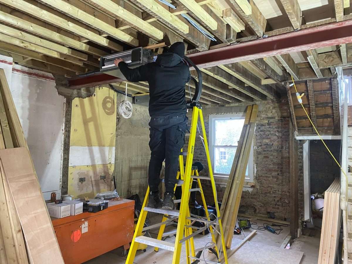 Fitter lifting a concealed ducted fan coil unit into an open ceiling void beside a steel beam
