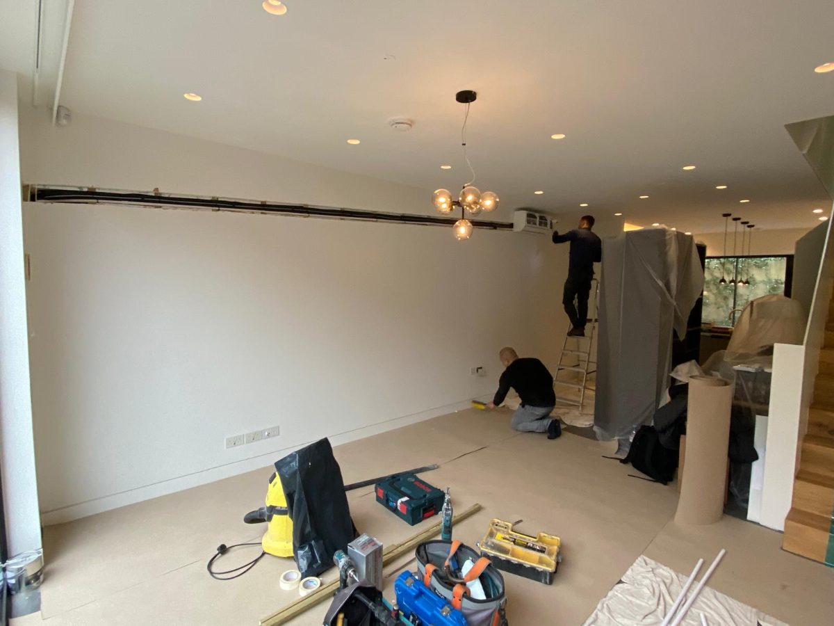Two fitters installing a wall-mounted split unit in a dust-sheeted living room, pipe run chased along the wall